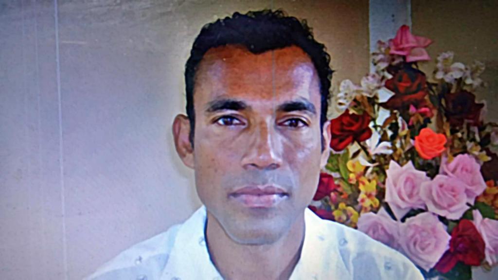 Retrato de un hombre de tez morena, con camisa blanca y flores en segundo plano.